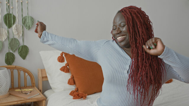 Young african american woman dancing on bed with arms raised and smiling, orange tassel pillow and bedside table visible inside a building; joy.