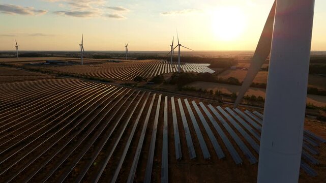 Aerial view of a vast sea of solar panels and spinning wind turbines bathed in golden hour sunset light at Chelveston Renewable Energy Park, UK