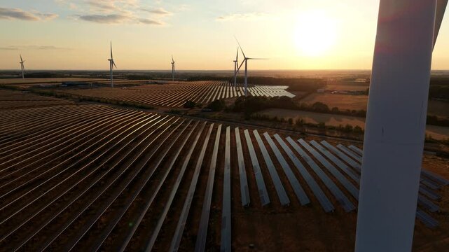 Aerial view of a vast sea of solar panels and spinning wind turbines bathed in golden hour sunset light at Chelveston Renewable Energy Park, UK