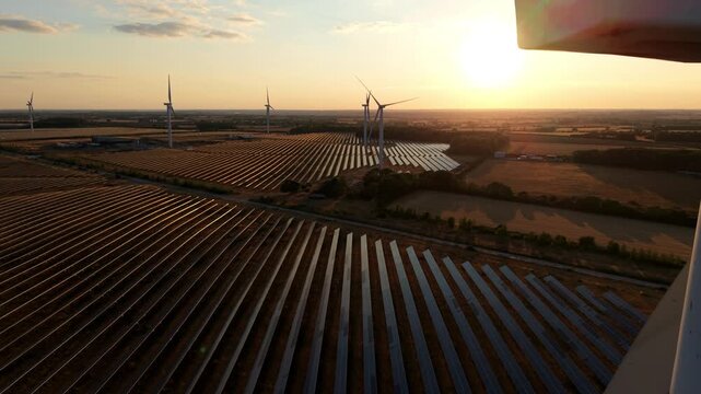 Aerial view of a vast sea of solar panels and spinning wind turbines bathed in golden hour sunset light at Chelveston Renewable Energy Park, UK