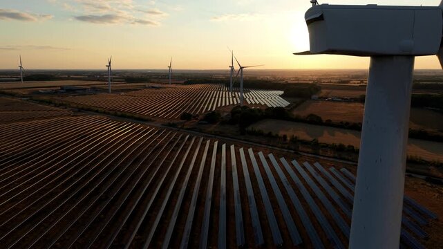 Aerial wide shot of hybrid solar and wind renewable energy farm glowing at golden hour sunset in Northampton UK