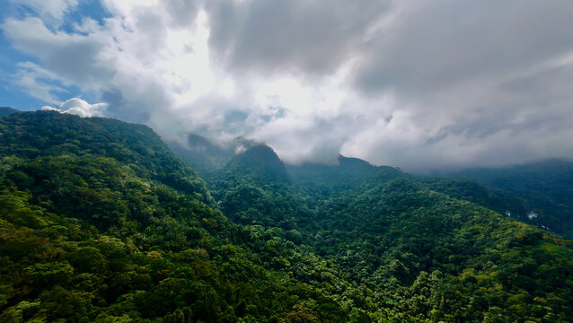 A vast green mountain range under a dramatic cloudy sky. Dense tropical forest covers the rolling hills as low clouds drift through the valleys.