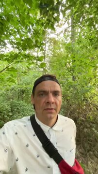 adult man walks forward along narrow dirt trail through dense deciduous woodland, captured from chest height selfie viewpoint, he wears backward black cap, white