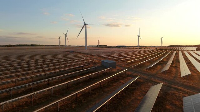 Forward aerial drone flight skimming over vast photovoltaic solar panel tables toward turning wind turbines silhouetted against a golden hour sunset sky.