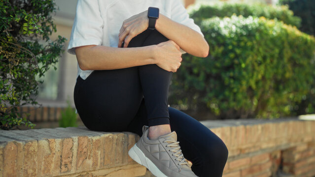 Woman in sportswear sits on a brick wall by a street, holding and rubbing her knee, legs crossed, wearing black leggings, grey sneakers and a smartwatch; calm.