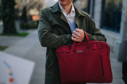 A businessman carries a red briefcase while standing on a city sidewalk. He wears a green coat and white shirt and holds the bag across his body as he pauses outdoors.