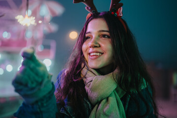 Naklejka premium Young woman in a scarf and antler headband smiles while holding a lit sparkler. A festive winter market scene with colorful lights and friends enjoying the holiday evening.