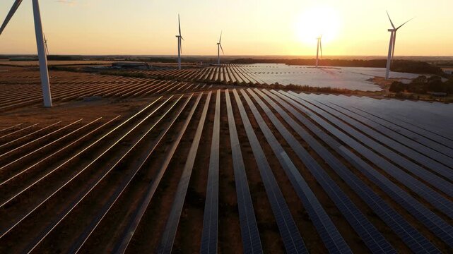 Forward aerial drone flight skimming over vast photovoltaic solar panel tables toward turning wind turbines silhouetted against a golden hour sunset sky.