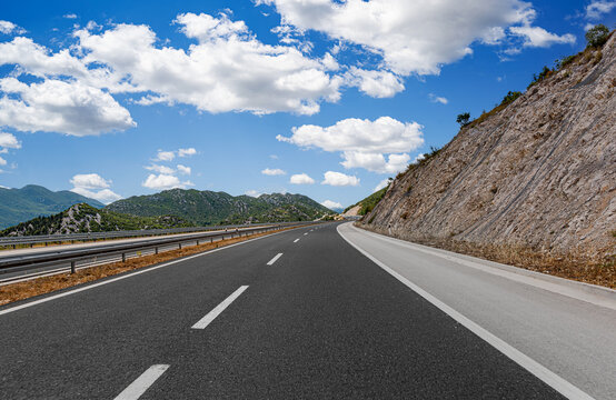 A paved highway winds through a scenic mountain landscape under a bright blue sky filled with white clouds.