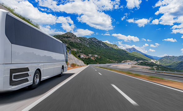 A modern white tour bus travels along a scenic highway through a mountain landscape under a bright blue sky filled with clouds.