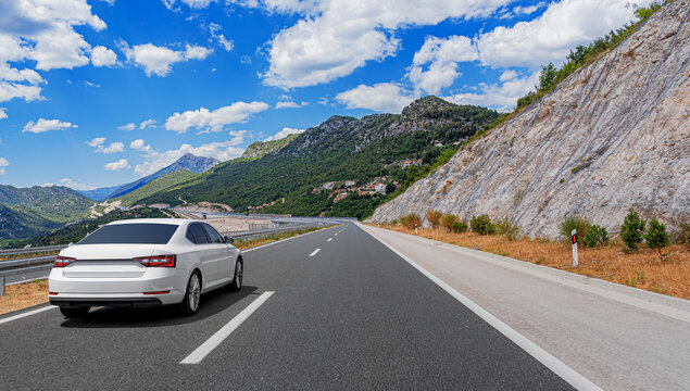 A white modern sedan drives along an open asphalt highway winding through scenic mountain landscapes under a bright blue sky with white clouds.