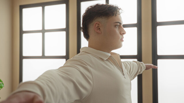 Young hispanic man with arms outstretched performing yoga t pose near large window in studio; serenity balance focus.