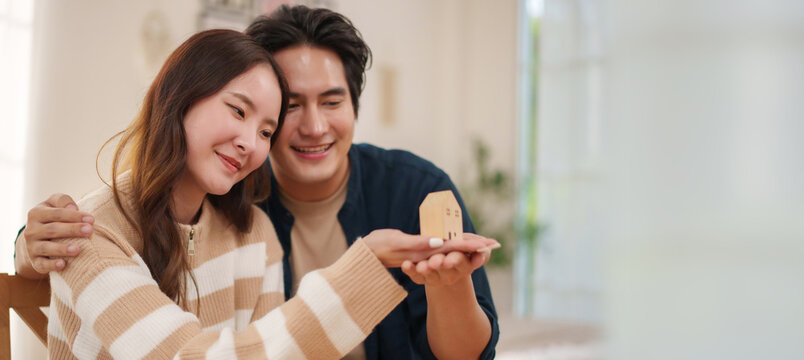 Happy Asian couple holding a miniature house while planning their future home together, representing property ownership.