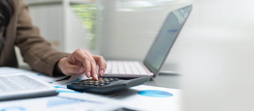 Businesswoman using a calculator while reviewing financial charts and reports beside a laptop, representing accounting, financial analysis, budgeting and business planning.