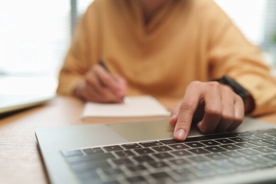 Woman browsing on laptop while writing notes in a notebook at a desk, representing multitasking, online research, business productivity, e-learning, planning and modern digital work.
