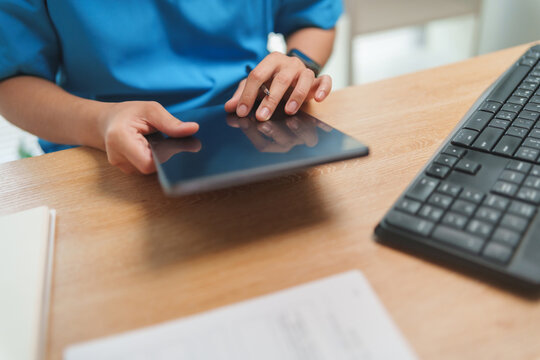 Doctor in blue scrub working with digital tablet at a medical desk, representing patient care, diagnosis and hospital technology.