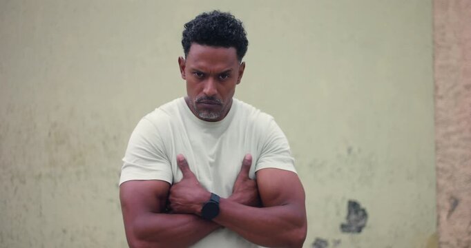 Man of African descent standing against textured wall with arms crossed, intense stare at viewer, serious confrontational expression, strong posture, tough and fearless attitude