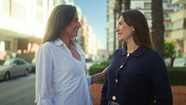 Two women pointing and smiling on a sunlit street by buildings and palm trees, one with earrings and a navy jacket; friendship joy.