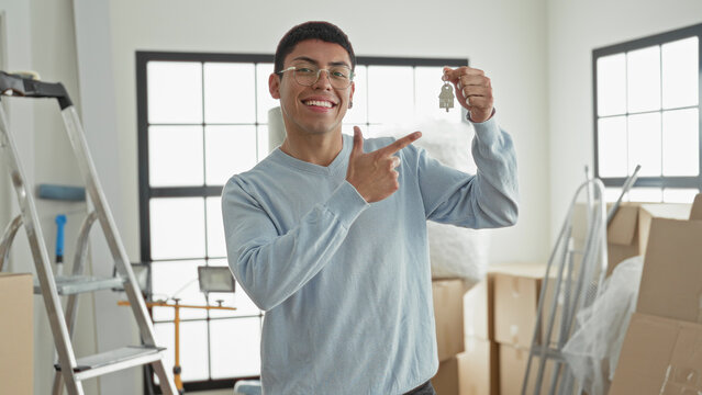 Man smiling points finger at key fob while holding keys among moving boxes and ladder in building; new home joy.