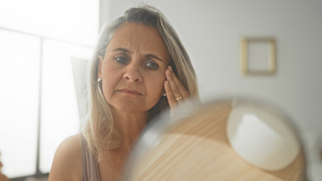 Woman touches cheek in round vanity mirror, hand on face inspecting wrinkles and forehead lines while seated at a mirror in studio; aging concern.