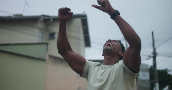 Man of African descent clenching fists and raising arms in triumph, looking up with powerful expression of victory, strength, resilience, achievement, and determination