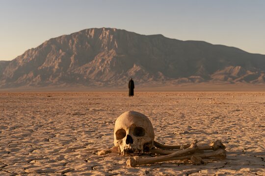 Human skull and bones on a dry desert landscape with a mysterious hooded figure standing in the background