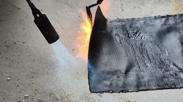 Detailed slow-motion footage of an open flame torch melting a bituminous waterproofing membrane on a flat roof, while a worker manually smooths the edge.