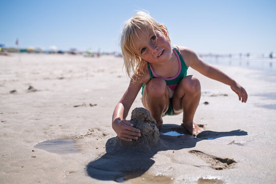 girl in green swimsuit sitting on sandy beach building sandcastles by ocean