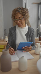 Fototapeta premium Woman with curly hair and glasses writing with a pencil while holding a tablet at a pottery studio workspace; concentration creativity.
