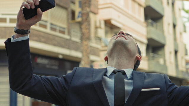 Man in suit raises smartphone aloft on street, holding phone above head while gazing upward; confidence ambition drive.