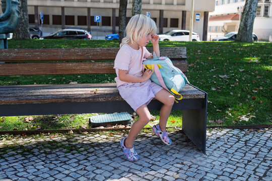 Sad little girl sitting alone on park bench with backpack