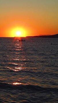 Sunset casts vibrant orange and yellow colors over the ocean, with boats gently bobbing on the water's surface, creating a serene evening atmosphere