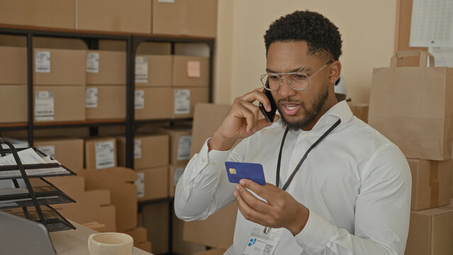 Man examining creditcard while speaking on smartphone to ear, holding card near face and scanning parcel labels in a storage building; concentration.