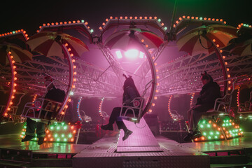 Three friends sit on a neon-lit swing ride at a nighttime carnival. The amusement ride glows with colorful lights and suspended seats as it rotates. © qunica.com
