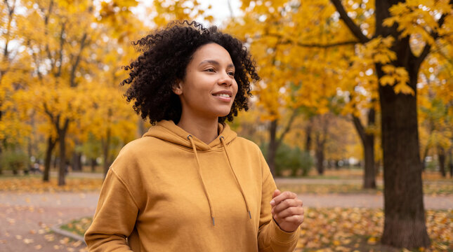 Young Afro-Latina woman walks briskly in a city park in a mustard hoodie, relaxed autumn mood, for active lifestyle social posts and fall marketing.