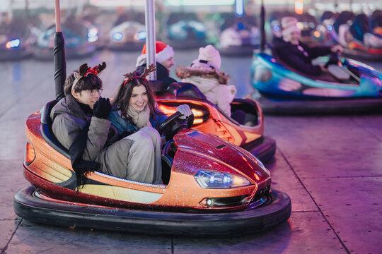 Young couple wearing reindeer antlers ride a glittering bumper car at a festive fairground. They smile and drive together among colorful lights and other bumper cars.