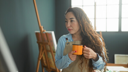 Woman holding orange mug and studying a canvas on an easel in a sunlit studio, wearing an apron  quiet contemplation. © Krakenimages.com