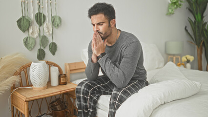 Fototapeta premium Man seated on bed with hands clasped to face and head bowed in a building bedroom; anxiety reflection.