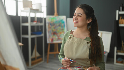 Woman painter holding a paint palette and brush, hand visible and smiling while looking at a canvas in studio  creative calm. © Krakenimages.com