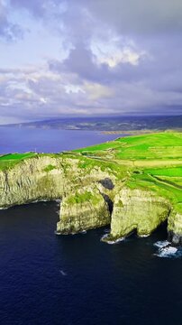 Sheer cliffs and rocky promontories along Sao Miguel coast, Azores, Portugal. Coastal drone footage of rugged Atlantic shoreline beneath farmland across green island hills