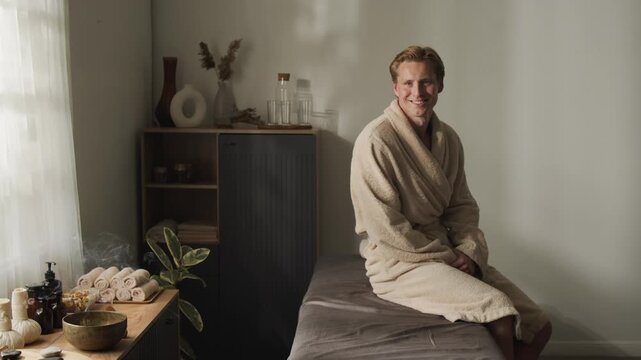 Portrait of smiling Caucasian young adult man sitting on massage bed in spa salon. Footage ideal for promoting wellness retreats, luxury body treatment services, and male self care campaigns