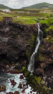 Coastal waterfall flowing from green cliff directly into ocean, Azores islands aerial view with Atlantic waves below