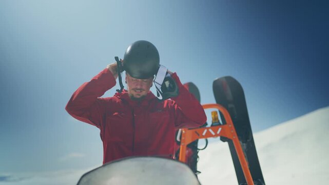 A man in a red jacket secures his ski helmet, preparing for winter sports in a snowy mountain landscape.