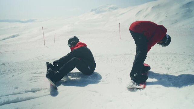 Two snowboarders are seen preparing their gear on a sunny, snow-covered mountain. They adjust their bindings before heading down the slopes.