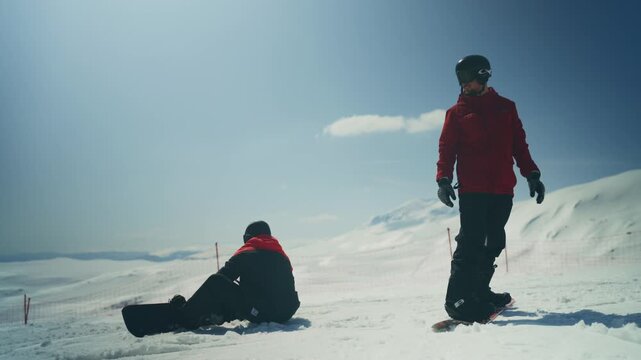 Two snowboarders relax and take in the panoramic views on a bright, snowy mountain slope. They are enjoying a beautiful day for winter sports and outdoor adventure.