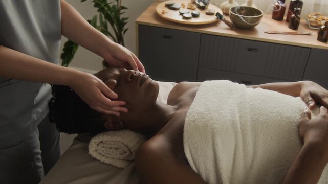 Caucasian masseuse performing facial massage on young adult African American woman lying on spa table. Footage works for wellness clinic campaigns, skincare ads, and beauty salon promotion