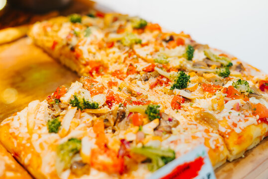 A vegetable pizza slice is placed on a wooden board, showcasing a mix of cheese, tomatoes, mushrooms, and broccoli. The pizza has a golden crust and appears freshly made, inviting viewers to enjoy