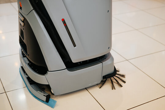 A robot floor cleaner is actively cleaning a tiled floor surface. The device features rotating brushes and is positioned at an angle as it works