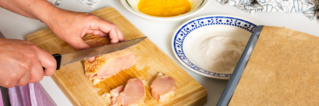 A woman cuts chicken on a wooden board with a knife. Nearby, there is a bowl of beaten eggs and a plate with flour, all set for making deep-fried chicken nuggets, banner