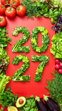 A festive flat lay arrangement featuring the numbers 2027 made from sliced greens, herbs, vegetables, and lettuce on a red board. A Christmas-themed concept for healthy, nutritious eating
